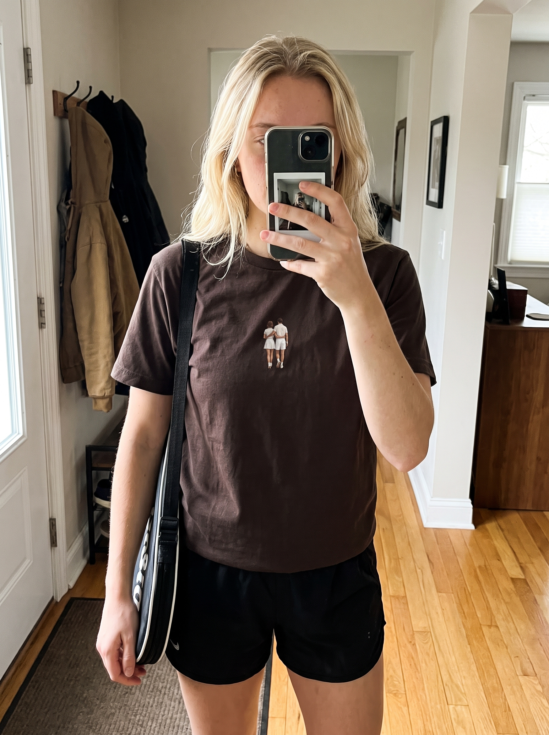 A woman takes a mirror selfie in a home hallway, wearing a dark brown crewneck t-shirt with a small graphic of two figures on the chest, paired with black athletic shorts and carrying a black shoulder bag.