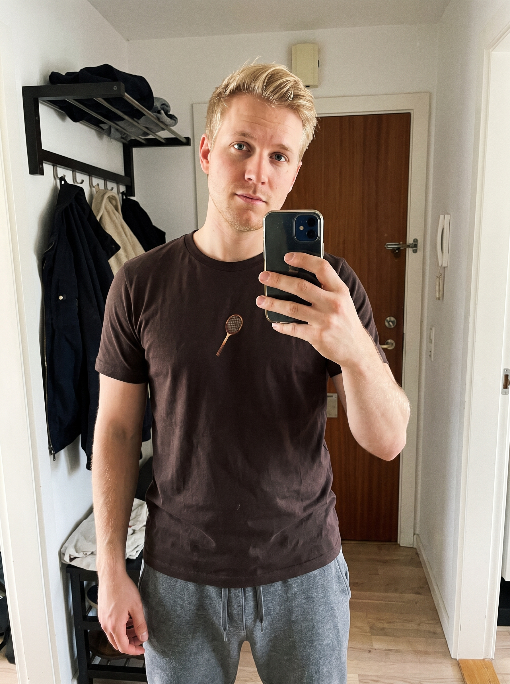 A man taking a mirror selfie in a hallway, wearing a dark brown crew-neck t-shirt with a small graphic of a spoon on the chest and gray sweatpants.