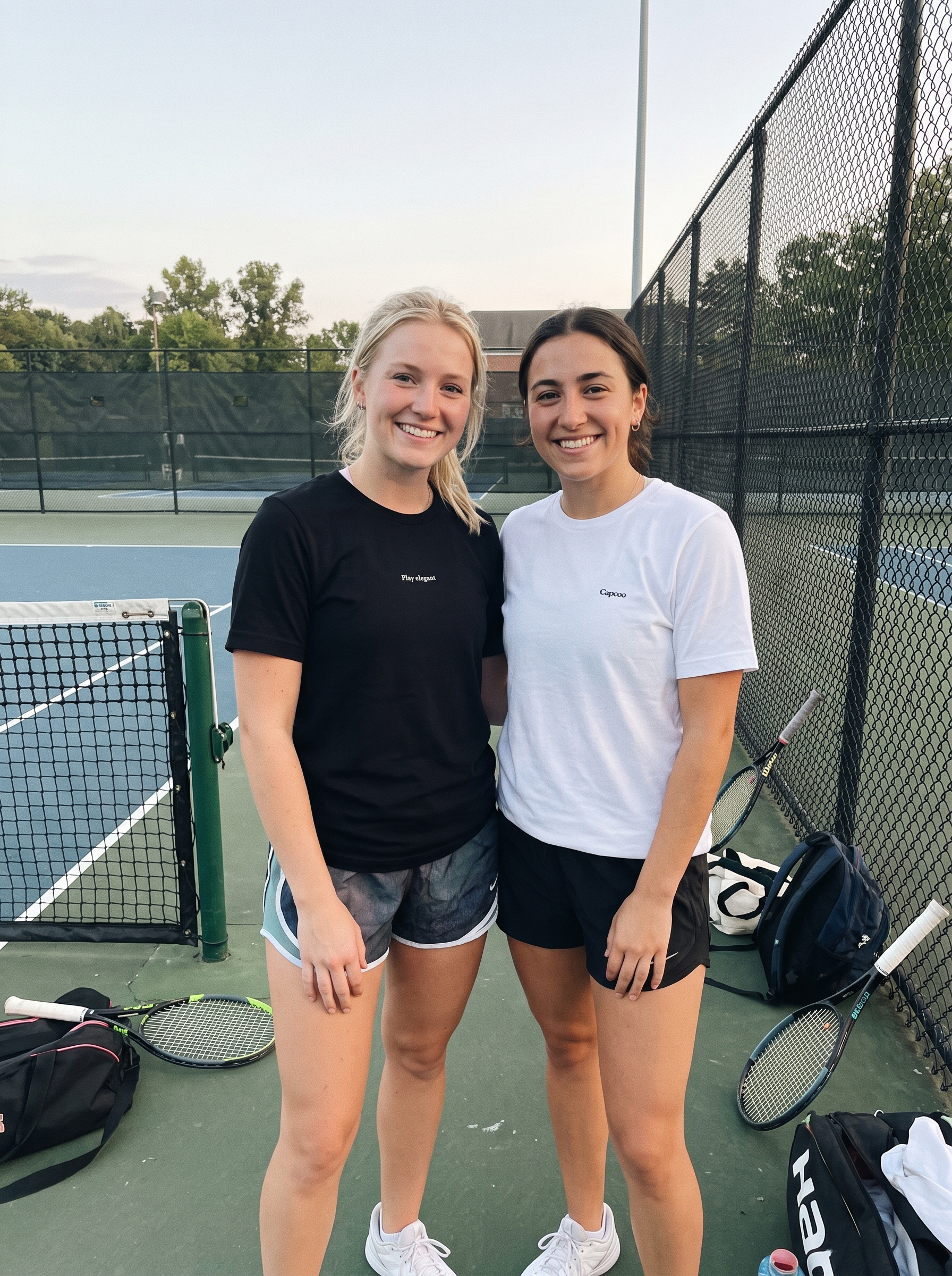 Two young women smiling and posing together on an outdoor tennis court. The woman on the left wears a black t-shirt and denim shorts, while the woman on the right wears a white t-shirt and black shorts.