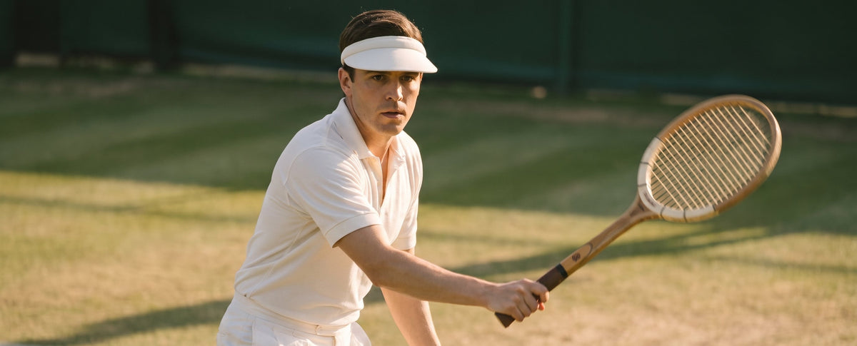 A man on a grass tennis court wearing a white tennis outfit, white visor, and holding a wooden tennis racket.