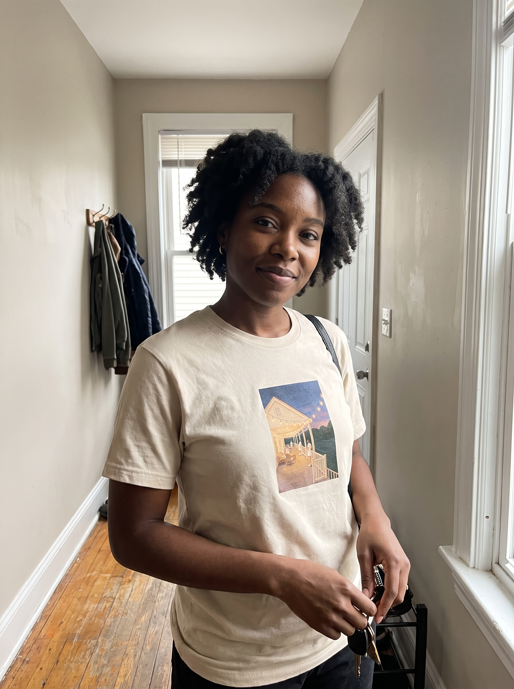 A woman with curly black hair stands in a hallway wearing a cream-colored t-shirt with a graphic print of a house, holding keys.
