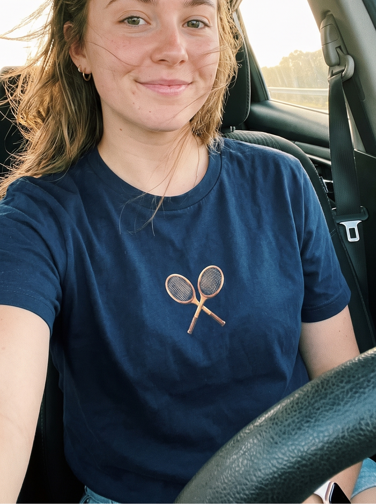 A woman sitting in the driver's seat of a car, wearing a navy blue t-shirt with a small graphic of two crossed tennis rackets on the chest.