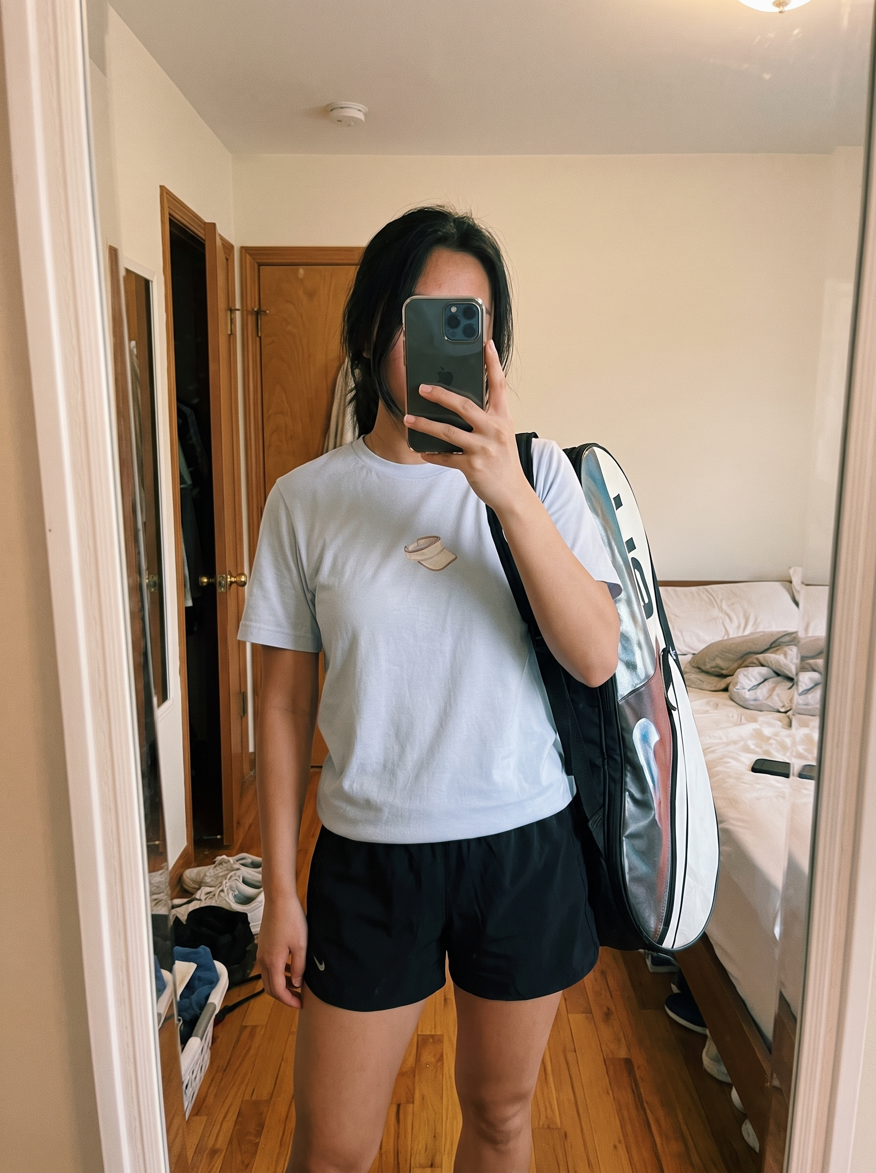 A person taking a mirror selfie in a bedroom, wearing a light gray t-shirt with a small graphic on the chest and black athletic shorts, while carrying a large tennis racket bag over their shoulder.